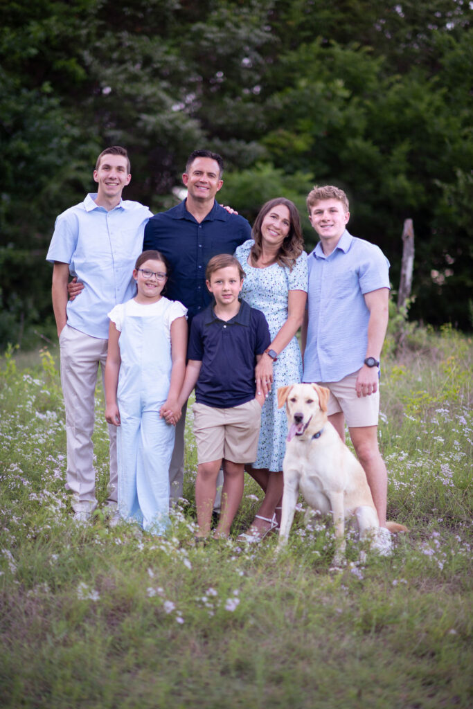 The Tripple family smiling together in a wildflower field, with parents, three children, one teenager, and a yellow Labrador retriever.