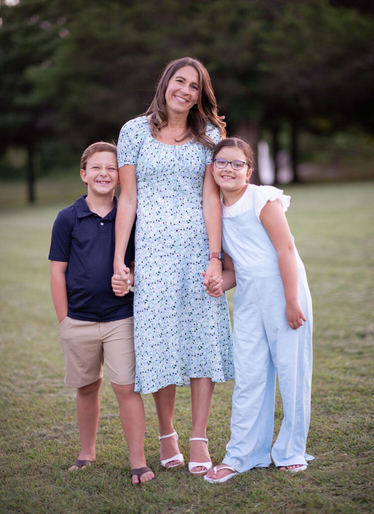 Michele Tripple standing outside holding hands with her two smiling children on a grassy field.