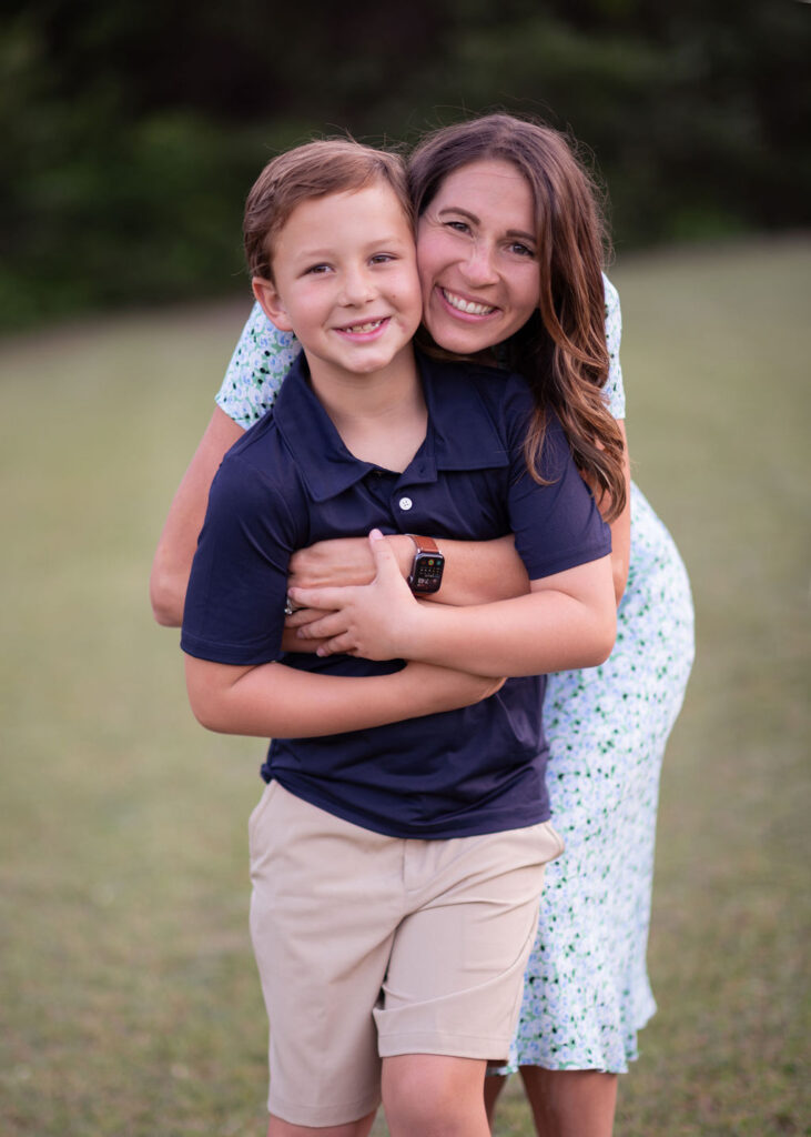 Michele Tripple hugging her smiling young son outdoors in a grassy field, both looking at the camera with joy.